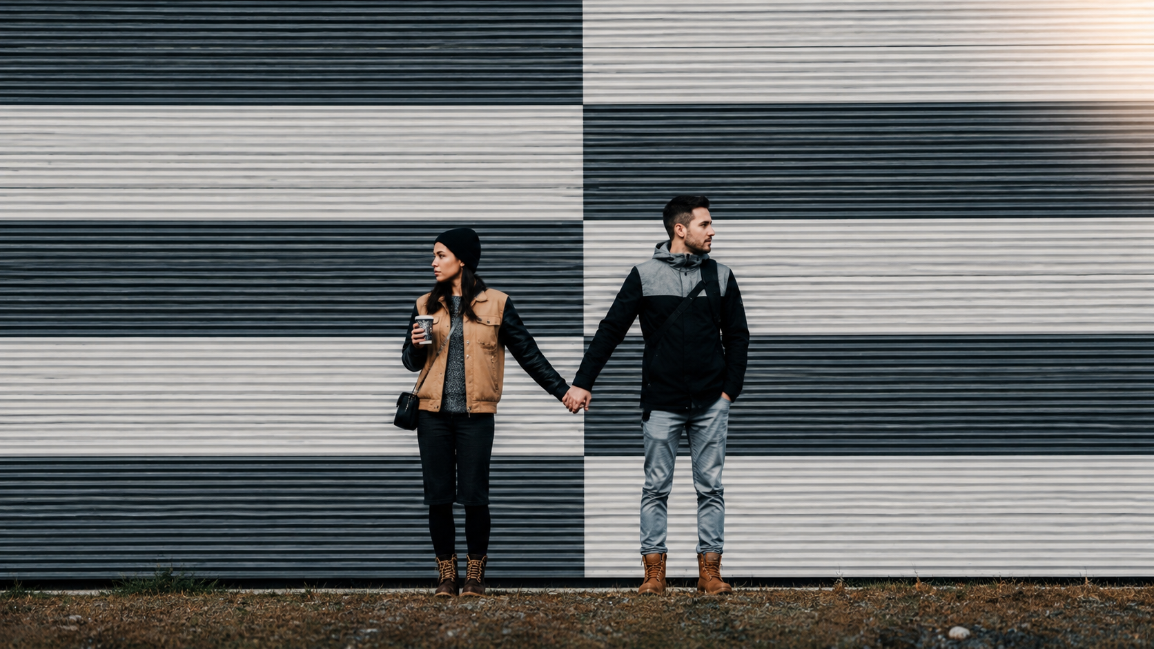 Contenuto articolo Una coppia si tiene per mano mentre guarda in direzioni opposte davanti a un muro con motivi geometrici bianchi e neri.